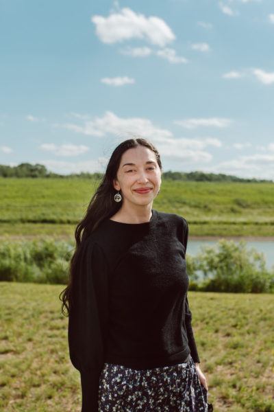 person with long black hair standing in front of water and field
